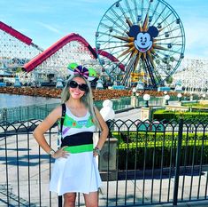 a woman standing in front of a fence with mickey mouse on it and a ferris wheel behind her