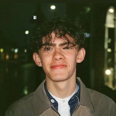 a young man in a trench coat looking at the camera with city lights behind him