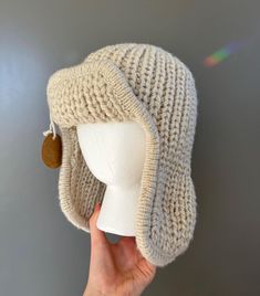 a hand holding up a white knitted hat with a brown button on it's brim