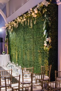 an indoor wedding setup with white flowers and greenery on the wall, along with gold chairs