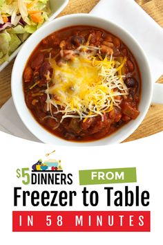 a white bowl filled with chili and cheese on top of a wooden table next to a salad