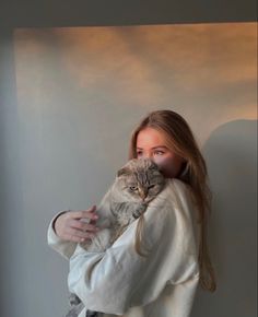 a woman holding a cat in her arms while standing next to a wall and looking at the camera