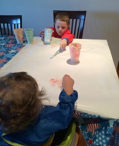 two young children sitting at a table with paper cups and paint on the table cloth