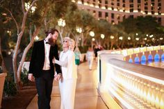 a man and woman walking down a sidewalk at night with lights on the buildings in the background