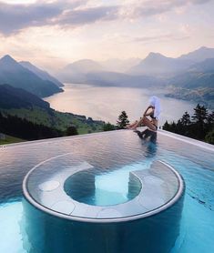 a woman sitting on the edge of a swimming pool with mountains and water in the background