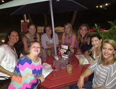 a group of women sitting at a table with drinks and papers under an umbrella outside