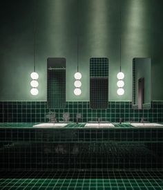 a green tiled bathroom with three mirrors and lights on the wall above the double sinks