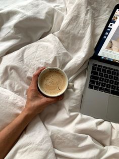 a person holding a cup of coffee in front of a laptop on top of a bed