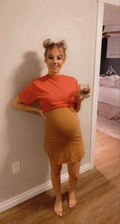 a woman in an orange shirt and brown skirt holding a glass of wine while standing next to a white wall