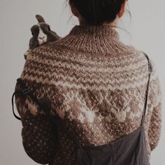 a woman wearing a brown sweater holding a cat in her back pocket while standing next to a white wall