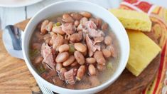 a white bowl filled with beans and ham on top of a wooden cutting board next to a piece of cornbread