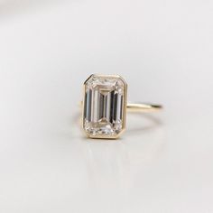 an emerald cut diamond sits on top of a white surface, with a gold band