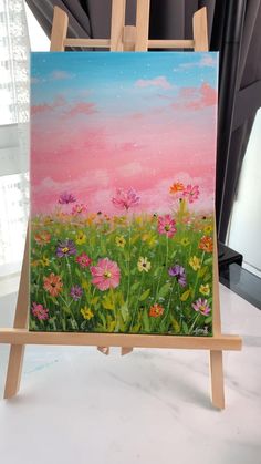 an easel with a painting on it that has flowers in the grass and pink sky