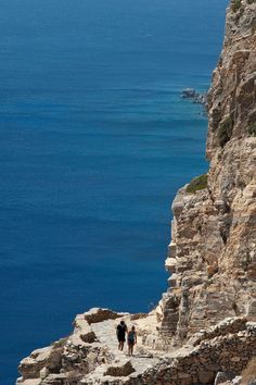 two people are walking up the side of a cliff by the ocean on a sunny day