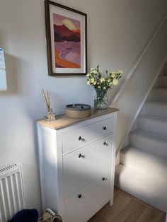 a white dresser sitting next to a stair case with flowers on it and a painting hanging above