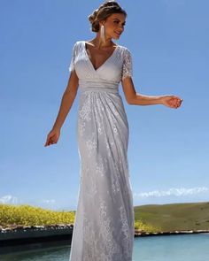 a woman in a long white dress standing on the beach with her hands out and looking off to the side