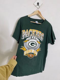 a green bay packers t - shirt hanging on a hanger next to a person's hand