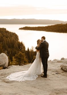 a bride and groom standing on top of a mountain looking at the lake in the distance