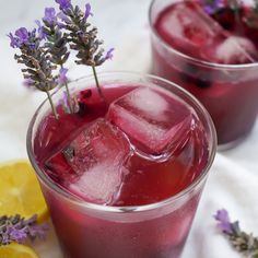 two glasses filled with ice and purple flowers on top of a white cloth next to lemon slices