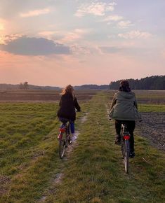 two people riding bikes down a dirt path in the middle of a field at sunset