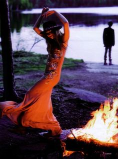 a woman in an orange dress sitting next to a campfire with a man standing behind her