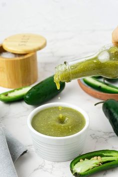 someone pouring green sauce into a small white bowl with cucumbers around it on a marble surface