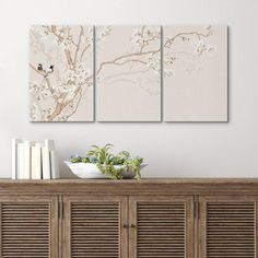 three paintings on the wall above a buffet with books and vases in front of it