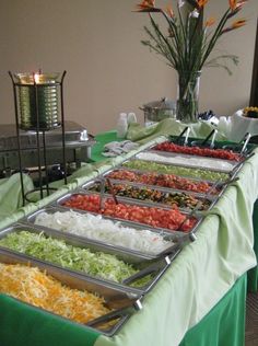 several trays of food are lined up on a long table with green and white cloth