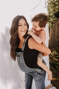 a woman holding a child in her arms and smiling at the camera while she is wearing overalls