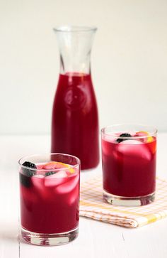 two glasses filled with red liquid next to a pitcher and a napkin on a table