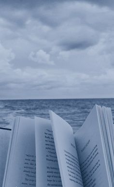 an open book sitting on top of a beach next to the ocean under a cloudy sky