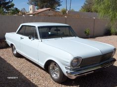 an old white car is parked on gravel in front of a fenced area with palm trees