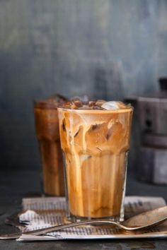 two iced drinks sitting on top of a table next to a spoon and fork with ice