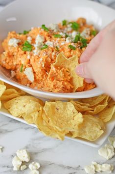 a person is dipping some chips into a white bowl with cheese and herbs on top