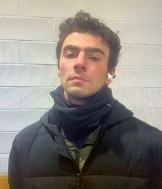 a man wearing a black jacket and scarf standing in front of a white brick wall