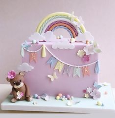 a birthday cake with a teddy bear and rainbow decoration on it's side, in front of a pink background