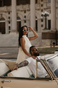 a man and woman sitting on the back of a convertible car in front of a building