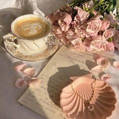 a cup of coffee and some pink flowers on a white table cloth next to an open book