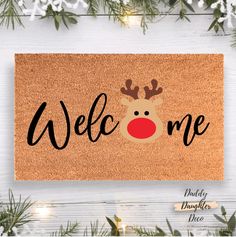 a door mat with the words welcome and a reindeer's nose on it, surrounded by pine branches