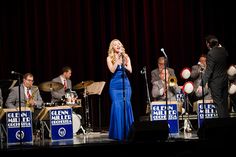 a woman in a blue dress sings into a microphone while other musicians play behind her