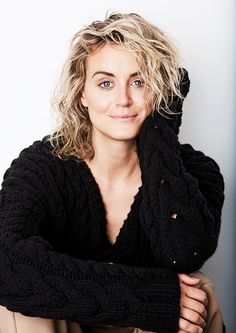 a woman with blonde hair wearing a black sweater posing for a photo in front of a white wall