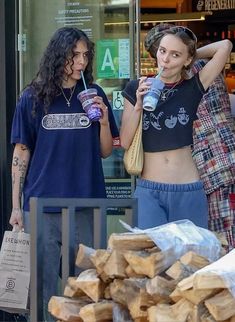 two young women standing in front of a store window drinking sodas and looking at their cell phones