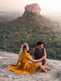 a man and woman sitting on top of a mountain next to each other with trees in the background