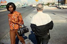 an older man standing next to a woman on the side of a road holding a purse