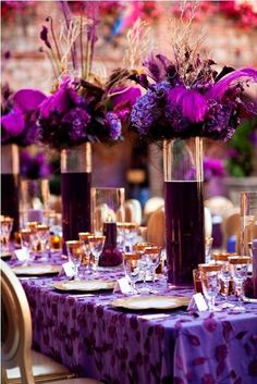 purple flowers are in tall vases with gold rimmed glasses and place settings on the table