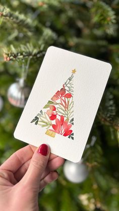 a hand holding up a card with a christmas tree design on it and ornaments in the background