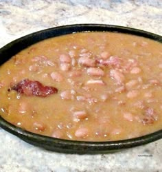 a black bowl filled with beans and bacon on top of a marble counter next to a white plate