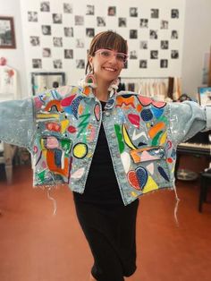 a woman wearing a jean jacket with colorful patches on the back and sides, standing in an art studio