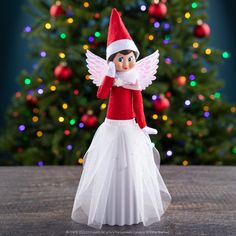 an elf doll sitting on top of a table next to a christmas tree with lights in the background