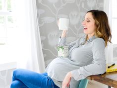 a pregnant woman sitting at a table with a cup in her hand and looking up